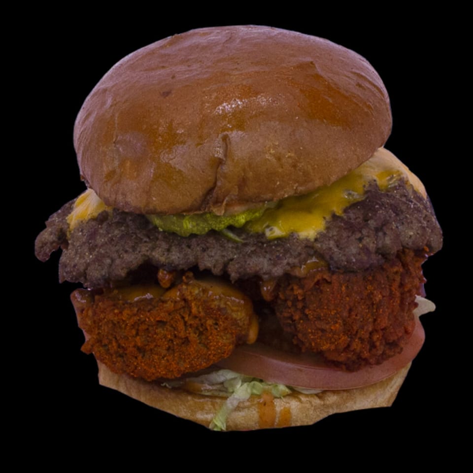 A large burger with a beef patty, cheese, guacamole, and two fried chicken pieces.