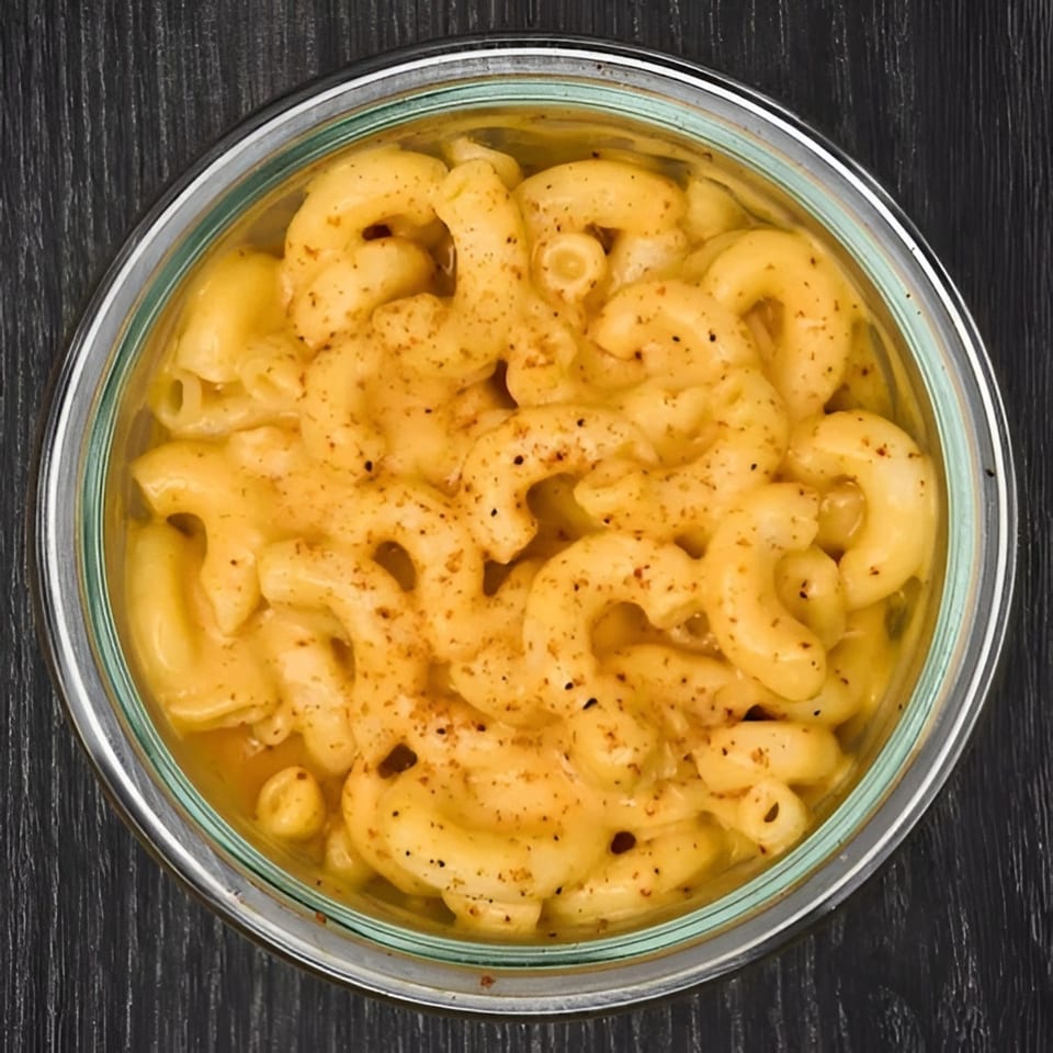 A glass bowl filled with creamy macaroni and cheese, topped with black pepper, on a dark wooden surface.