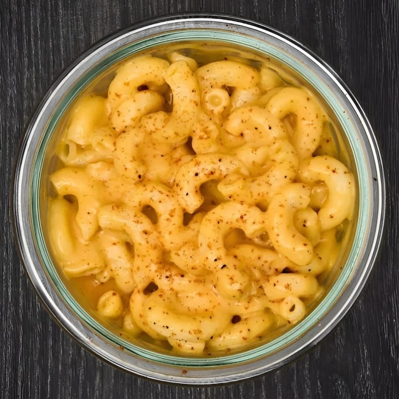 A glass bowl filled with creamy macaroni and cheese, topped with black pepper, on a dark wooden surface.